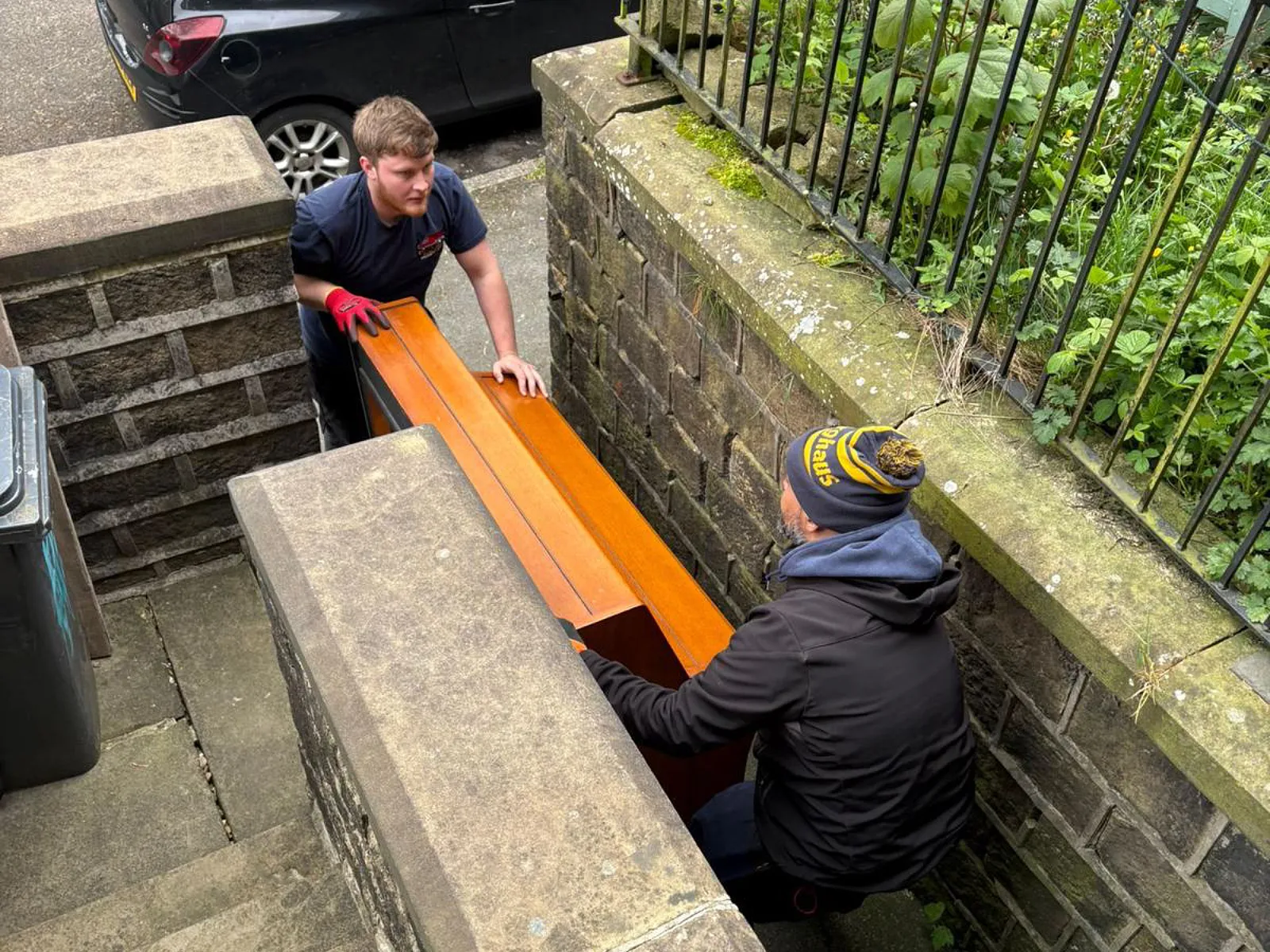 On The Move Removals two-person team carefully lowering a wrapped piano down outdoor stone steps during a specialist piano move in Huddersfield