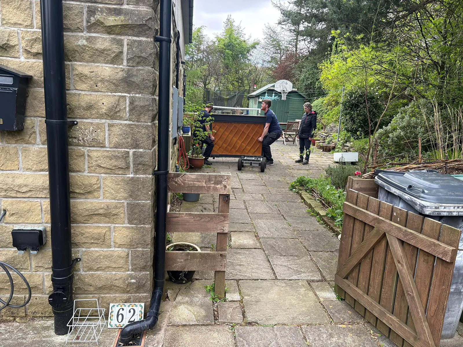 On The Move Removals two-person team walking a wrapped piano on a specialist dolly along a garden path during a Huddersfield piano move