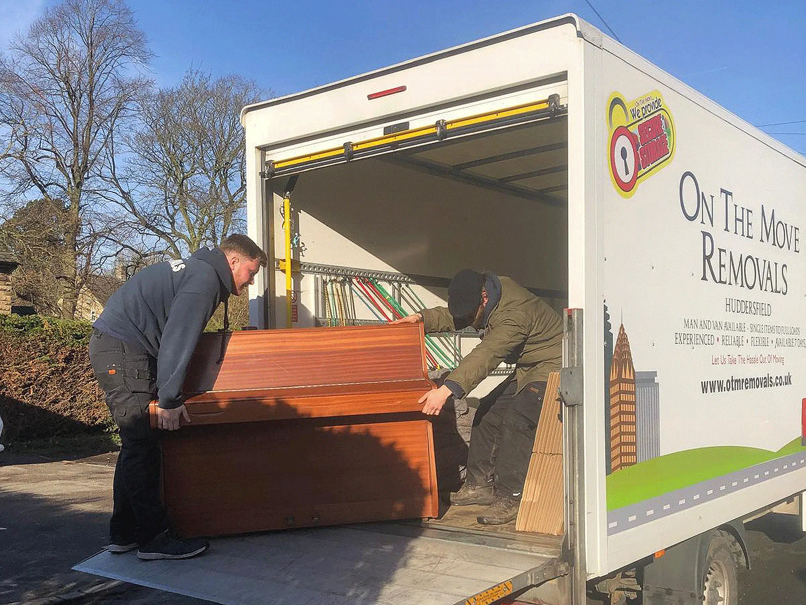 On The Move Removals two-person team unloading a wrapped upright piano off the back of a branded OTM van during a Huddersfield piano removal